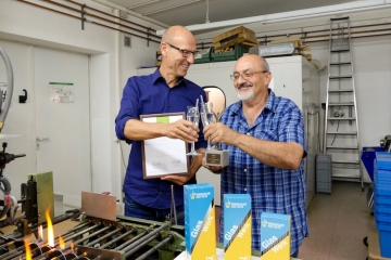 Geschäftsführer Axel Dostmann und Glasbläsermeister Ewald Kunkel sind stolz über den Award für das nachhaltige Produkt: Trinkhalme aus Glas GlasWerk. In einer kleinen Runde stoßen sie auf die Auszeichnung an. Im Hintergrund sieht man die Produktionsstätte der Trinkhalme.