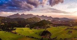 Panorama über Forggensee im Sommer bei warmen Temperaturen. Der Himmel ist bereits rosa gefärbt und die Wolken wirken violett.