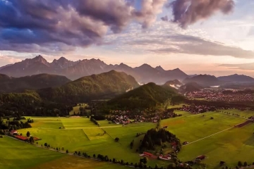 Panorama über Forggensee im Sommer bei warmen Temperaturen. Der Himmel ist bereits rosa gefärbt und die Wolken wirken violett.
